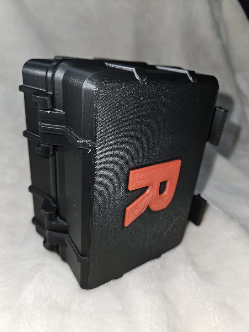 May include: A black, rectangular case with a textured surface and a bright orange letter "R" on the front. The case has a hinged lid and clasps. The overall design suggests a protective container.