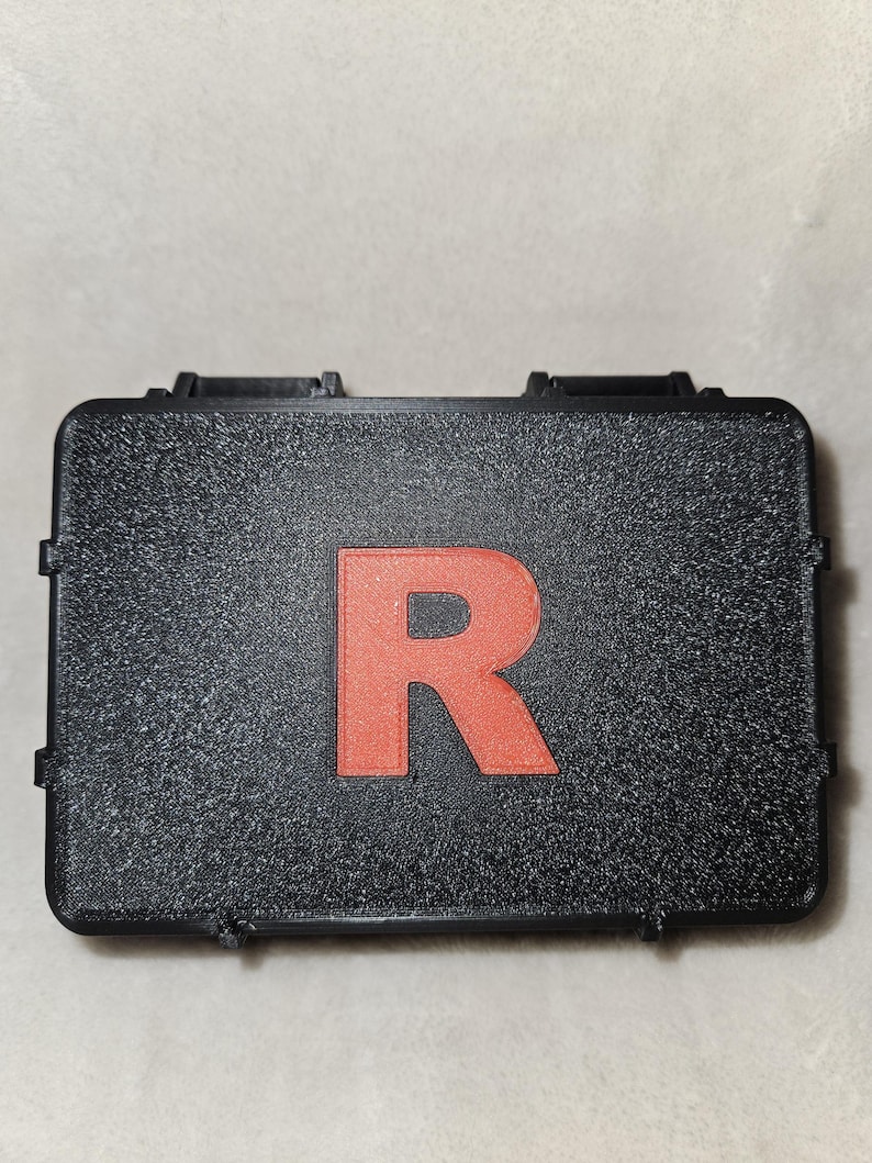 May include: A black rectangular case with a textured surface. A large, red letter "R" is centered on the lid. The case has two black clasps on the front.