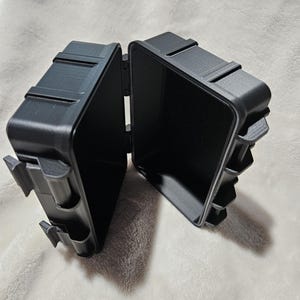 May include: A black, open, hinged box with a rectangular shape. The box is made of a matte material and has a textured surface. The box is open, revealing the empty interior. The box has a clasp on the side.