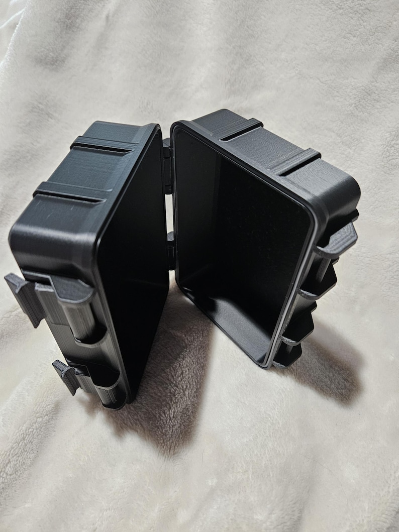 May include: A black, open, hinged box with a rectangular shape. The box is made of a matte material and has a textured surface. The box is open, revealing the empty interior. The box has a clasp on the side.