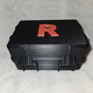 May include: Black rectangular case with a red letter "R" on the top. The case has a textured surface and a hinged lid with clasps. The case appears to be made of plastic.