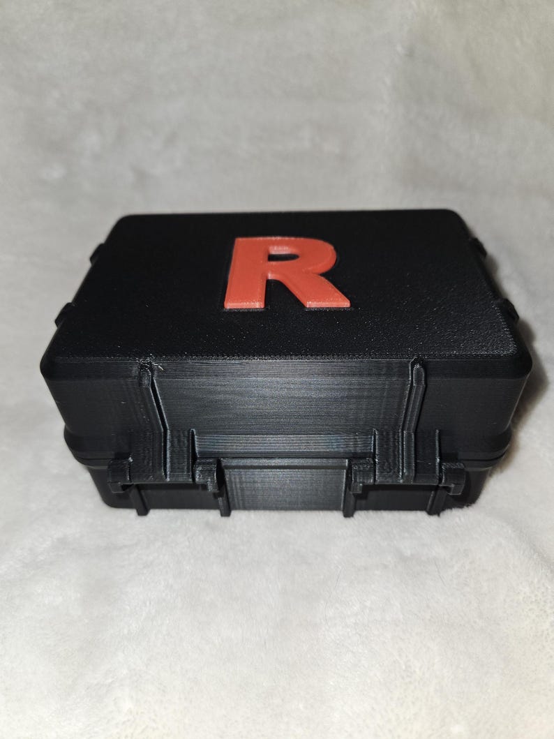 May include: Black rectangular case with a red letter "R" on the top. The case has a textured surface and a hinged lid with clasps. The case appears to be made of plastic.