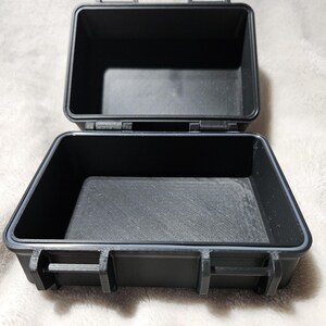 May include: An open, black, rectangular case with a hinged lid. The case is made of a durable material and features a gray rubber seal around the edges. The interior is empty, and the case is designed to protect its contents.