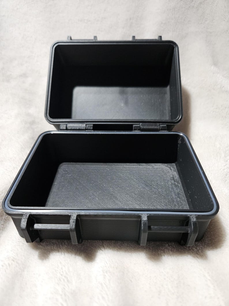 May include: An open, black, rectangular case with a hinged lid. The case is made of a durable material and features a gray rubber seal around the edges. The interior is empty, and the case is designed to protect its contents.
