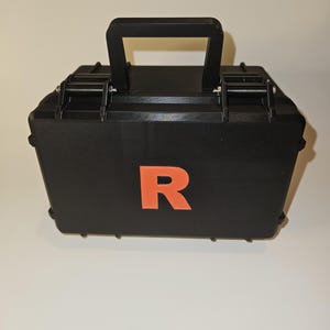 May include: A rectangular black hard-shell storage case featuring a top carrying handle, two secure locking latches, and a prominent orange letter R printed in the center of the front panel. The case has a textured finish and a rigid construction.