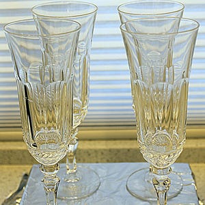 May include: Four clear champagne flutes with intricate etched designs. The glasses have a tall, slender shape with a wide base and a detailed stem. The flutes are arranged on a light-colored surface, suggesting a formal setting.