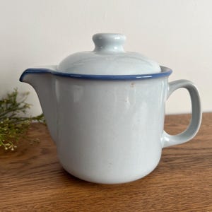 May include: A light blue ceramic teapot with a matching lid and a blue trim. The teapot has a curved handle and a spout for pouring. The teapot is sitting on a wooden surface.