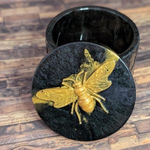 May include: A black resin container with a lid featuring a gold-coloured bee design. The bee is detailed with textured wings and body. The container is round and has a glossy finish, suitable for storing small items. The background is a wooden surface.