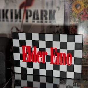 May include: A rectangular sign featuring a black and white chequered pattern across the entire surface. The words "Elder Emo" are displayed in bold, raised, bright red 3D lettering centred horizontally and vertically on the front of the sign.