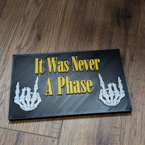 May include: A rectangular black plaque featuring the yellow raised text "It Was Never A Phase" centred on the surface. Two white skeletal hands are positioned on the left and right sides of the text, each displaying the rock and roll hand gesture.