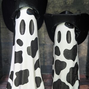 May include: Two white ghost figurines with black cow-like spots and black cowboy hats. The ghost figures have oval-shaped eyes and a curved mouth. The hats have a wide brim and a tall crown. The background is a dark grey.