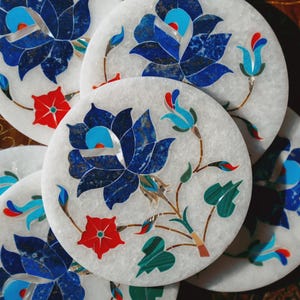May include: A collection of white marble coasters with intricate floral designs. Each coaster features a large blue flower with detailed petals, complemented by red and turquoise accents. The coasters are round and appear to be handcrafted.