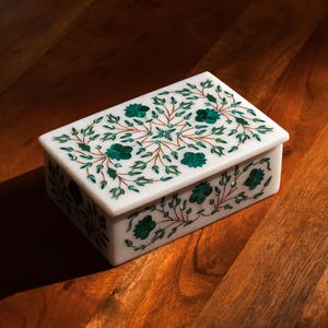 May include: A rectangular white marble box with a lid, adorned with a floral inlay design. The inlay features green floral patterns and brown stems. The box is set on a wooden surface.