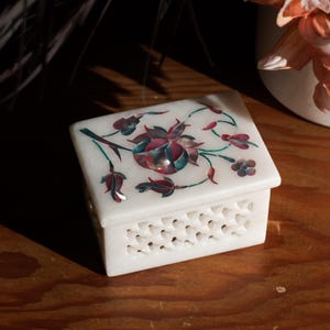May include: A white marble jewelry box with a floral inlay design in shades of red, teal, and green. The box has a square shape with a decorative pierced pattern around the sides. The box is sitting on a wooden surface.