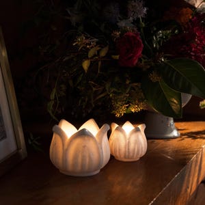May include: Two white ceramic lotus flower candle holders with lit candles inside, sitting on a wooden surface. The candle holders are in the foreground, with a dark background of flowers and a picture frame.