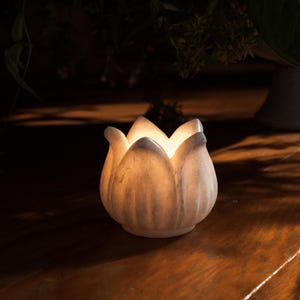 May include: A white, lotus-shaped candle holder with a soft, glowing light emanating from within. The ceramic piece sits on a dark wooden surface, creating a warm and inviting ambiance. The candle holder is a decorative item.