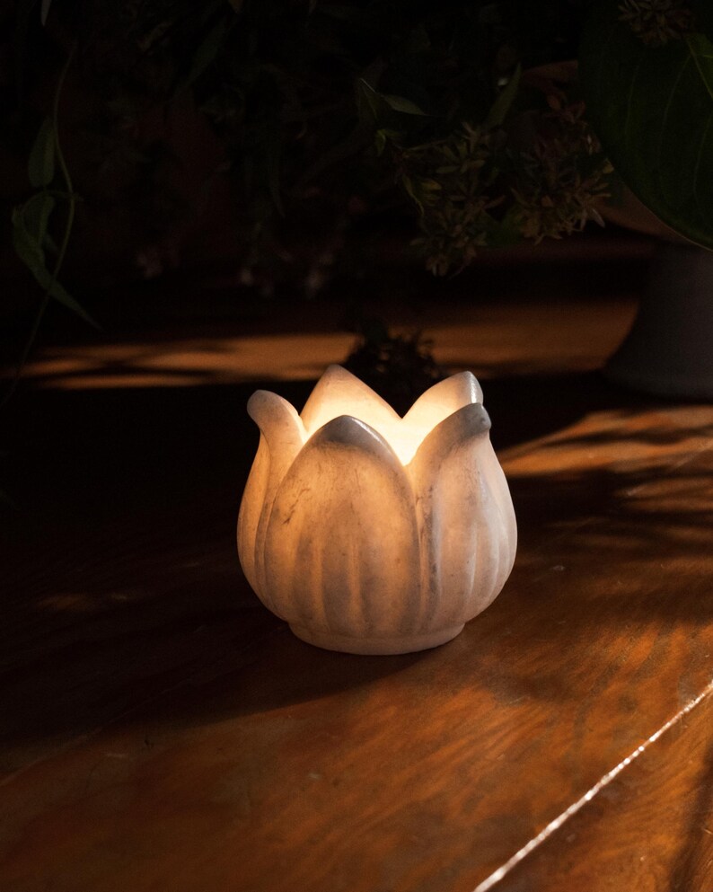 May include: A white, lotus-shaped candle holder with a soft, glowing light emanating from within. The ceramic piece sits on a dark wooden surface, creating a warm and inviting ambiance. The candle holder is a decorative item.