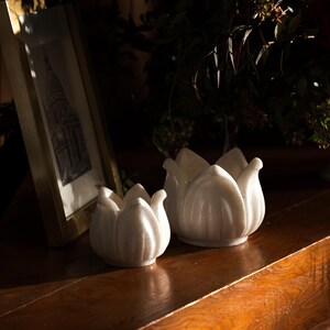 May include: Two white ceramic lotus flower candle holders on a wooden surface. The candle holders are different sizes and have a glossy finish. A framed picture and greenery are in the background.