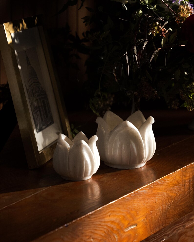 May include: Two white ceramic lotus flower candle holders on a wooden surface. The candle holders are different sizes and have a glossy finish. A framed picture and greenery are in the background.
