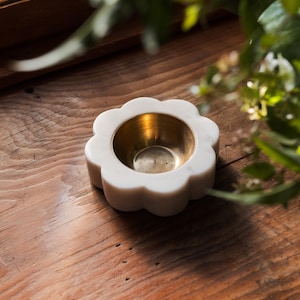 May include: A white marble flower-shaped bowl with a gold-colored interior. The bowl sits on a wooden surface, with greenery in the background. The bowl is likely used for decorative purposes.