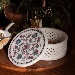 May include: White marble jewelry box with an oval lid featuring a floral inlay design in shades of pink, blue, and green. The box has a lattice-like pattern around the sides and sits on a wooden tray.