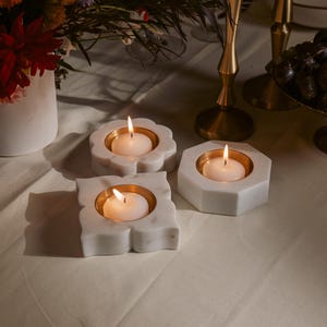 May include: Three white marble candle holders with gold-colored interiors, each holding a lit white candle. The candle holders have different shapes: a flower, a square, and a hexagon. A vase of flowers and brass candlesticks are in the background.