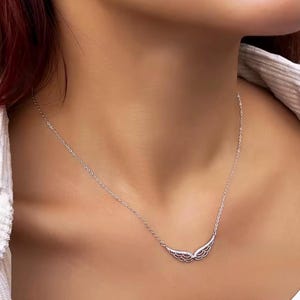 Op de afbeelding: Een zilveren ketting met een vleugelvormige hanger. De hanger heeft twee gedetailleerde vleugels, elk met ingewikkelde veerdetaillering. De ketting wordt getoond op de hals van een persoon, waarbij het ontwerp van de sieraden wordt benadrukt.