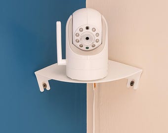 May include: A white security camera with a rounded top and multiple lenses is mounted on a white corner shelf. The camera has a single antenna and is positioned against a blue and beige wall.