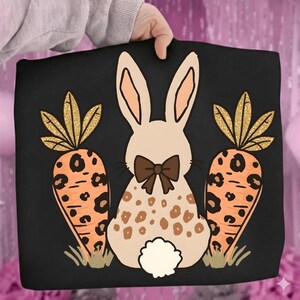 May include: Black tote bag featuring a bunny with a brown bow tie and a spotted pattern, flanked by two orange carrots with gold glitter tops. The design is set against a black background.