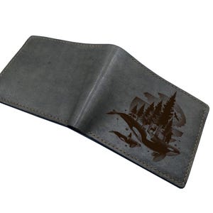 Może przedstawiać: Szary skórzany portfel z brązową ilustracją orek i sceną leśną. Portfel jest otwarty, ukazując wnętrze. Krawędzie są przeszyte brązową nicią. Projekt znajduje się po prawej stronie.