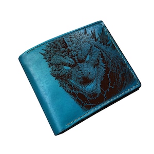 May include: A teal leather wallet featuring a detailed illustration of a monster's face with glowing blue eyes. The design is in dark blue and black, with visible stitching around the edges. The wallet is rectangular and appears to be bi-fold.