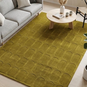 May include: A large rectangular area rug in a deep olive green colour featuring a repeating pattern of raised, rounded rectangular shapes across its entire surface. The rug is positioned on a light-coloured wooden floor in a living room setting.