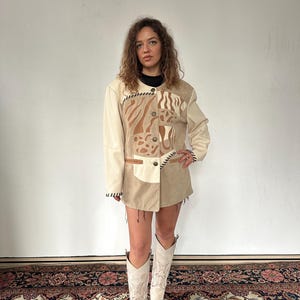 Peut inclure: Veste en cuir beige et marron clair avec un motif animalier unique. La veste a un col rond et une fermeture boutonnée. Le modèle porte un haut noir et des bottes de cowboy beiges claires. La veste a une esthétique vintage.