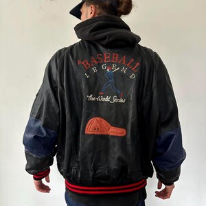 Puede incluir: Chaqueta de béisbol de cuero negro con detalles azules y rojos. La parte trasera presenta un diseño bordado con las palabras "BASEBALL LEGEND" y un gráfico de jugador de béisbol. También es visible un parche de gorra de béisbol.