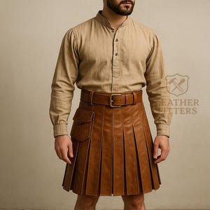 May include: A brown leather kilt with a pleated design, worn with a tan button-up shirt and a brown leather belt. The kilt features a pocket on the side. The shirt has a mandarin collar. The logo "Leather Fitters" is visible.
