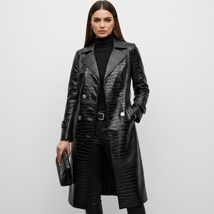 Può includere: Lungo trench coat nero in ecopelle con texture coccodrillo. Il cappotto presenta un design doppiopetto con bottoni argentati, una cintura e un colletto classico. Una pochette nera abbinata è tenuta nella mano sinistra.