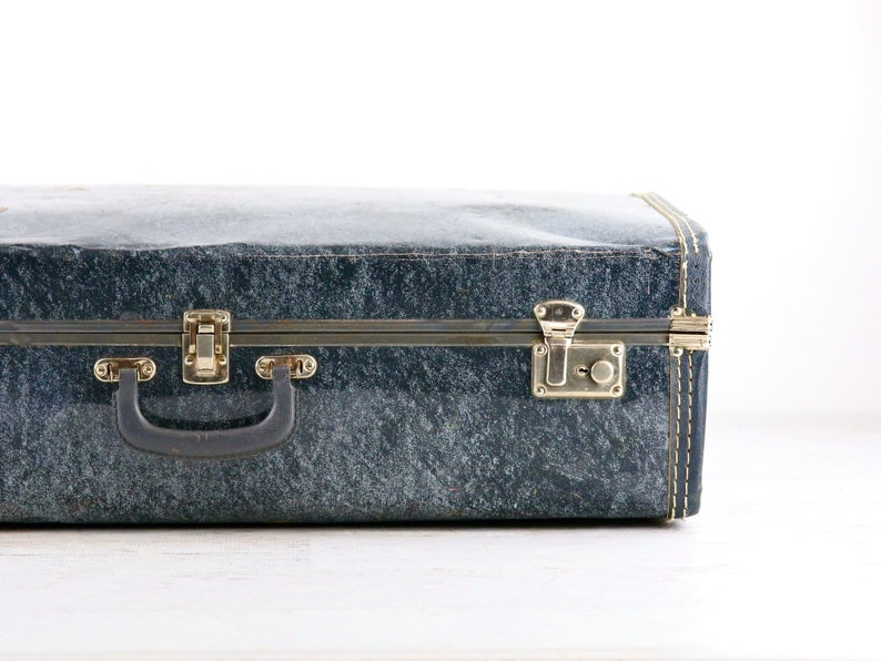 Op de afbeelding: Een vintage blauwe koffer met messing beslag en een zwarte handgreep. De koffer heeft een getextureerd oppervlak en is gesloten.