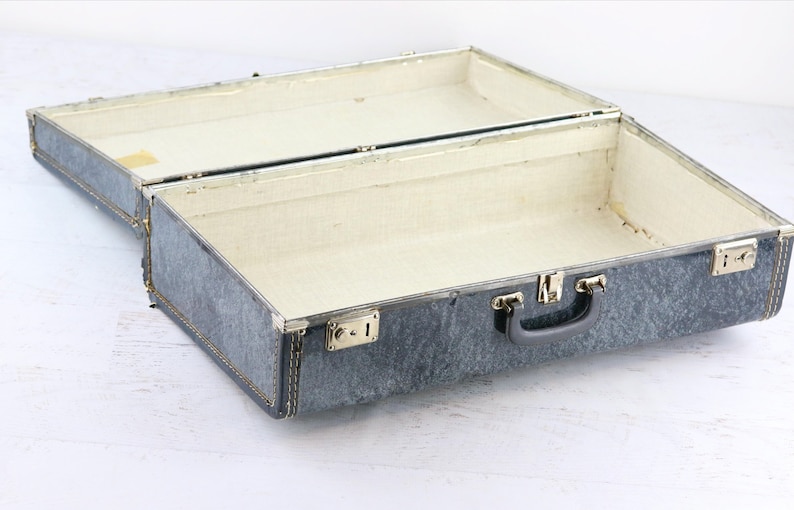 Op de afbeelding: Een vintage grijze koffer met een stoffen voering en een zwarte handgreep. De koffer is open en leeg.