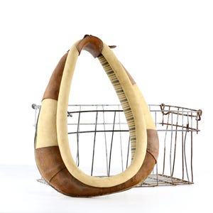 May include: A teardrop-shaped horse collar featuring alternating panels of smooth tan leather and textured cream-colored fabric. The collar is positioned in front of a rectangular wire mesh basket constructed from thin, weathered metal rods.