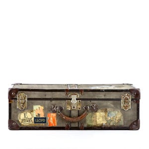 Peut inclure: Une valise rectangulaire grise avec des renforts d'angle en similicuir marron foncé et une poignée centrale. L'extérieur présente plusieurs étiquettes de voyage vintage, dont des autocollants avec le texte E LLOYD et LLOYD, et des fermoirs en métal.