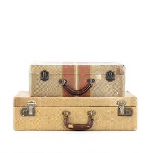 Peut inclure: Deux valises rectangulaires empilées verticalement. La valise supérieure est beige avec une bande verticale marron et jaune et une poignée foncée. La valise inférieure est beige clair avec un motif à chevrons et une poignée marron usée.