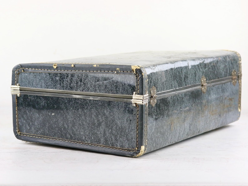 Op de afbeelding: Een vintage grijze koffer met een metalen sluiting en een versleten, getextureerd oppervlak.