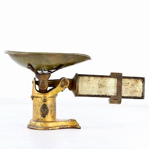 May include: Antique brass scale with a weathered appearance. The scale features a round, shallow bowl and a rectangular measurement guide. The base and supporting structure are gold-toned, with visible signs of age and use.