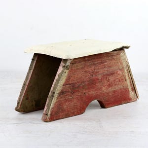 May include: A small, rustic wooden stool with a red base and a cream-colored top. The stool has a simple, A-frame design and shows signs of wear, giving it a vintage appearance. The dimensions are approximately 12 inches wide, 10 inches deep, and 10 inches tall.