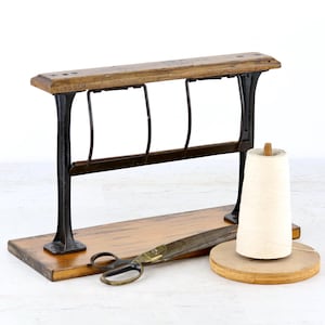 Peut inclure: Un support de bobines en bois et métal noir avec une base rectangulaire, doté d'un cadre vertical à trois séparateurs en fil métallique. À côté se trouvent une paire de ciseaux en métal et un cône de fil blanc sur une base ronde.