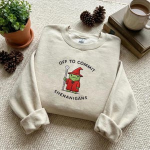 以下が含まれることがあります： 「OFF TO COMMIT SHENANIGANS」の文字と、魔法使いの帽子とローブを着た漫画のカエルが描かれたクリーム色のスウェットシャツ。カエルは杖を持っています。スウェットシャツは、テクスチャ加工された表面に折りたたまれています。