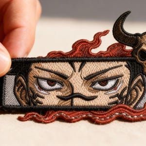 May include: Embroidered patch featuring a stylized face with intense eyes and a bull skull. The patch has a black border, tan and brown details, and red flame-like accents. The patch is held by a hand.