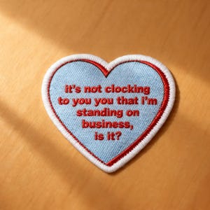 May include: Embroidered heart-shaped patch with a light blue center, white border, and red stitching. The patch features the phrase "It's not clocking to you that I'm standing on business, is it?" in red.