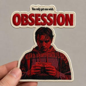 Op de afbeelding: Een geborduurde patch met een persoon in een rode trui met patroon die een kleine kaart vasthoudt. Boven de figuur staat de tekst "You only get one wish." en het woord "OBSESSION" in grote rode hoofdletters op een crèmekleurige achtergrond.