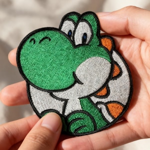 May include: Embroidered patch featuring a green dinosaur character with white, orange, and black accents. The patch has a circular shape and is held between two hands. The character has large eyes and a friendly expression.